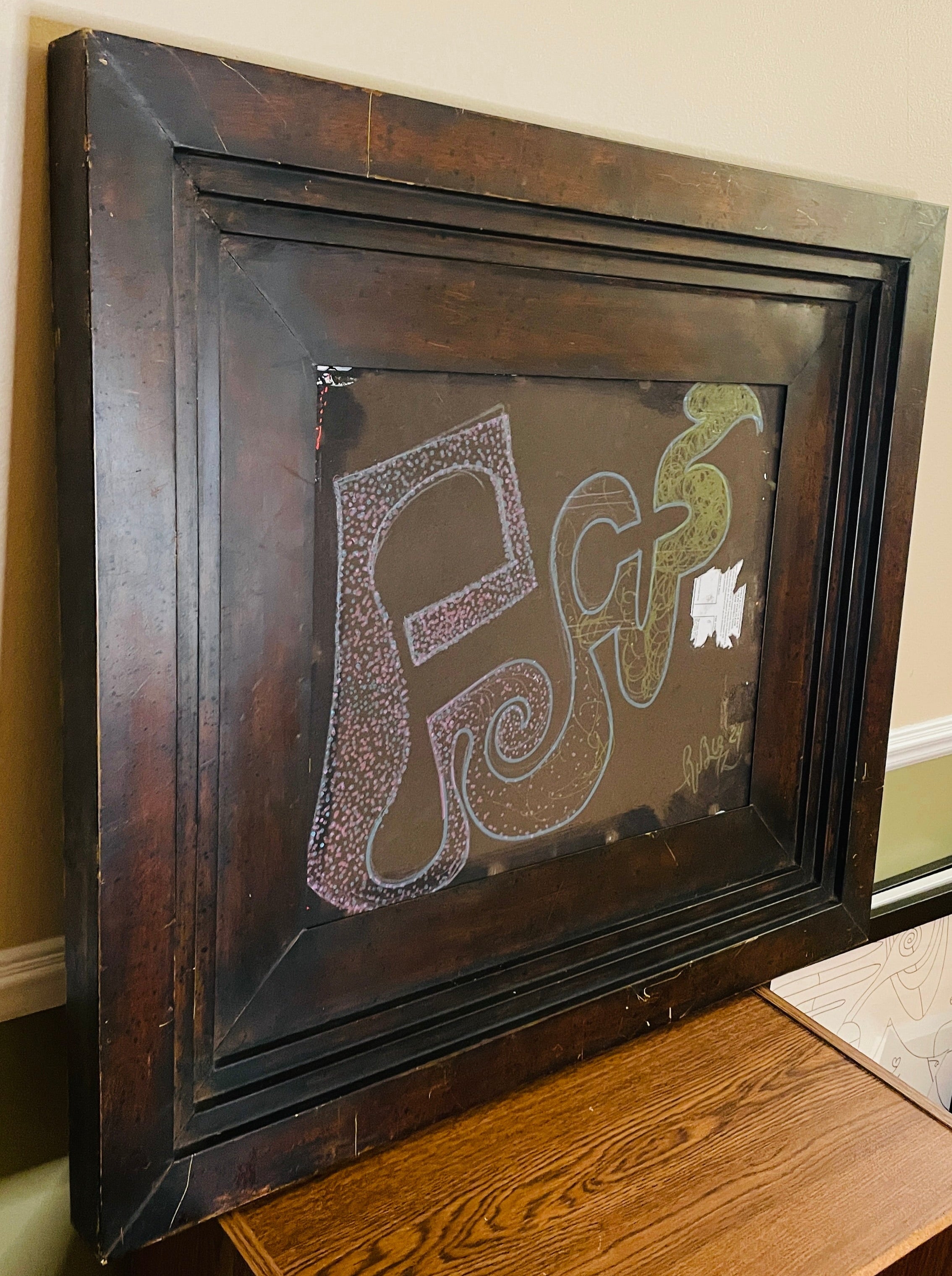 Abstract mixed-media assemblage in a deep, weathered dark wood frame. Pink, blue and green invented glyphs, dots, spirals and tangled lines appear on a brown field beside a torn fragment of printed paper.