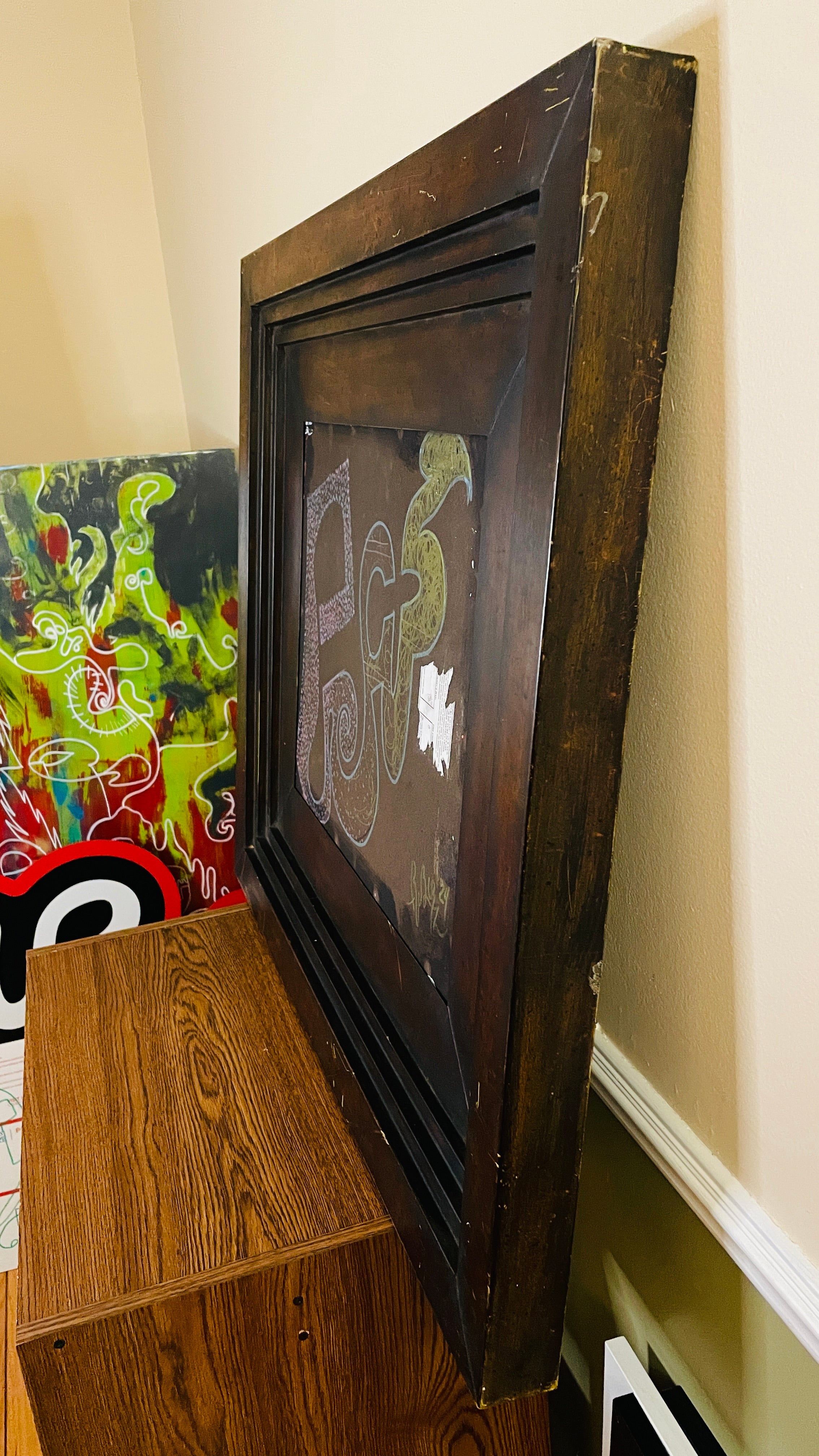 Abstract mixed-media assemblage in a deep, weathered dark wood frame. Pink, blue and green invented glyphs, dots, spirals and tangled lines appear on a brown field beside a torn fragment of printed paper.