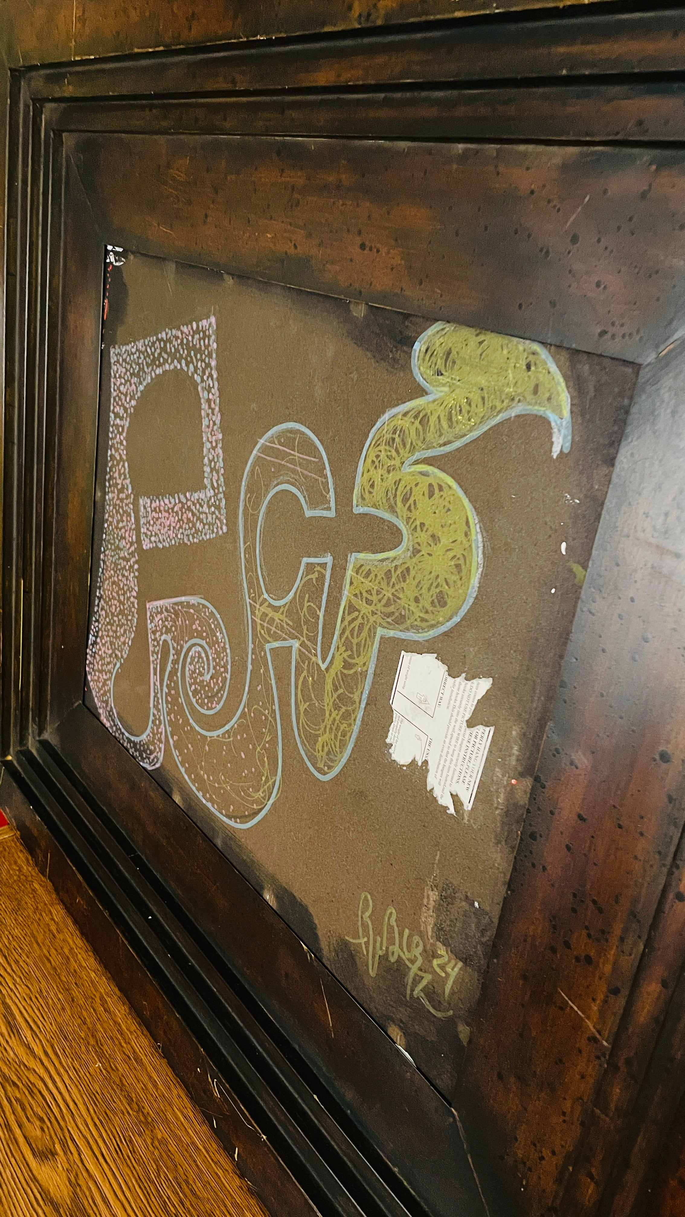 Abstract mixed-media assemblage in a deep, weathered dark wood frame. Pink, blue and green invented glyphs, dots, spirals and tangled lines appear on a brown field beside a torn fragment of printed paper.