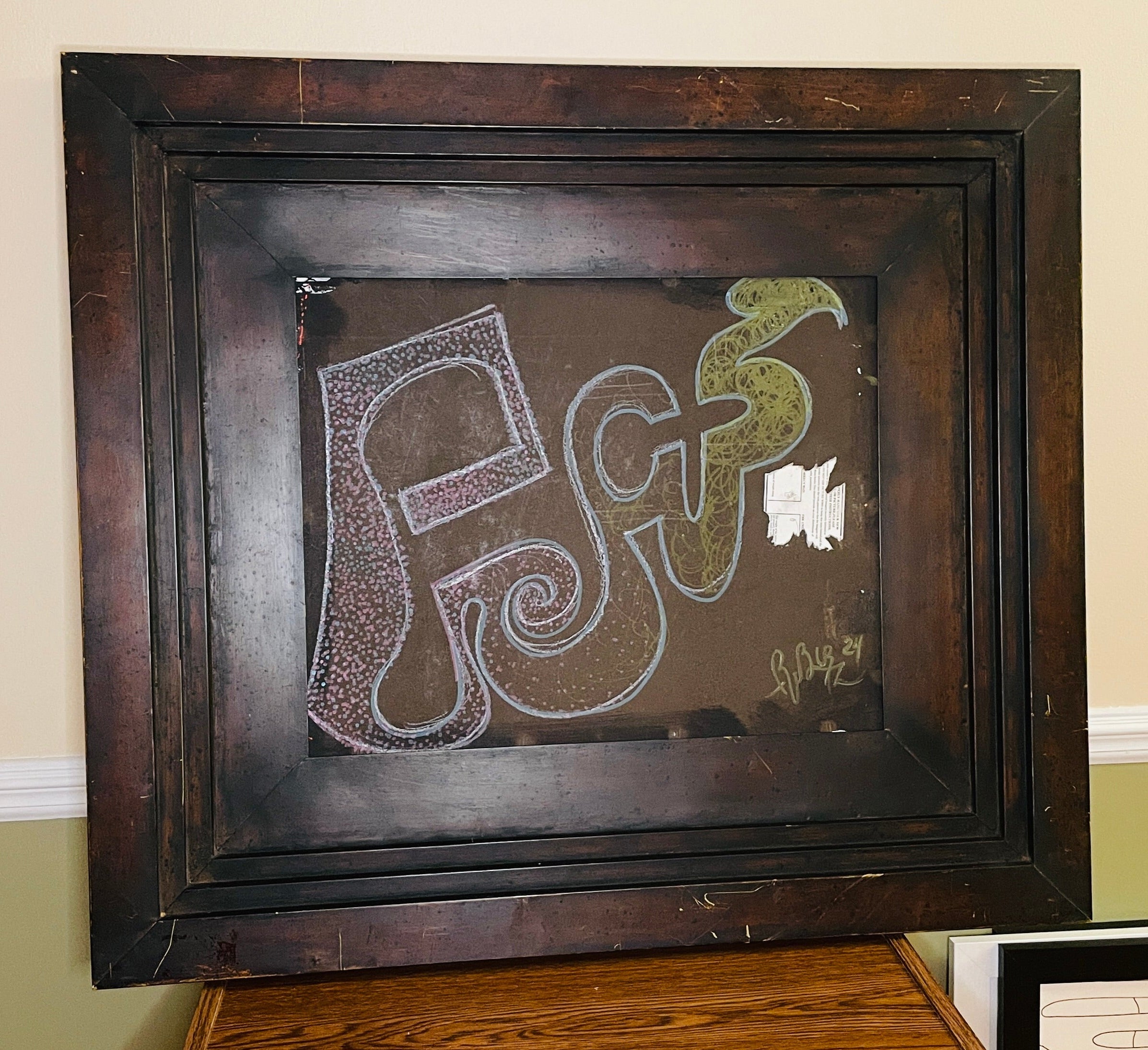Abstract mixed-media assemblage in a deep, weathered dark wood frame. Pink, blue and green invented glyphs, dots, spirals and tangled lines appear on a brown field beside a torn fragment of printed paper.